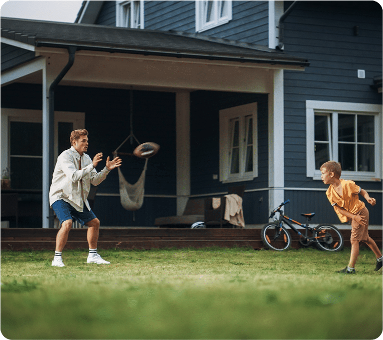 A child throws a football to a man in a backyard, with a blue house and bicycle visible in the background.
