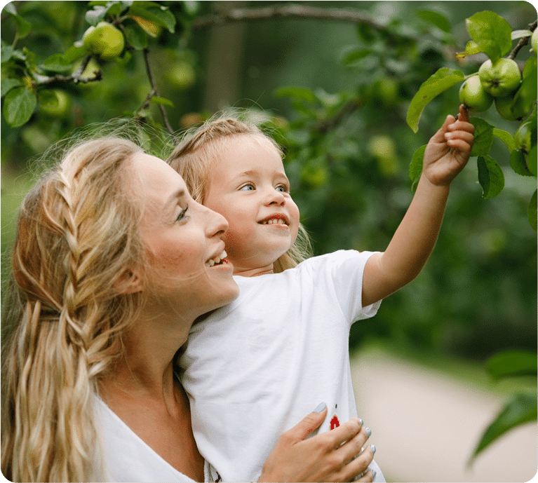 A mother holds her child in an orchard, pointing at green apples on a tree. The scene conveys joy and connection in nature.