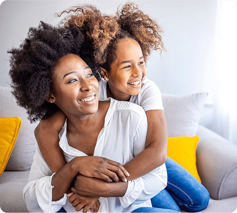 Two women with curly hair embrace on a light sofa, with bright sunlight filtering through sheer curtains, and a yellow pillow nearby.