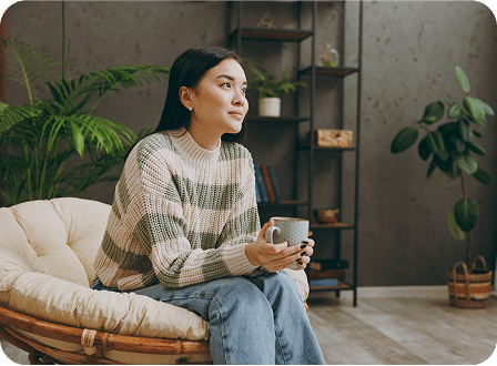 A person sits comfortably on a chair holding a mug, surrounded by greenery and neutral-toned decor in a cozy indoor space.